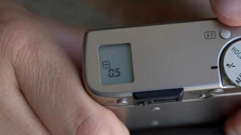 A close-up of a person's hand holding a camera, showing the top display screen with a reading of "-0.5" and part of the camera's control dial.