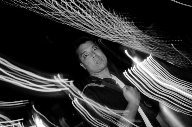 A man stands with a surprised expression among swirling, streaked lights at night, captured in black and white with motion blur.