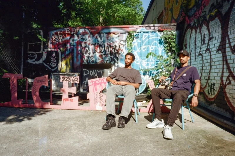Two people sit on chairs in front of a wall covered in colorful graffiti. They are outdoors on a sunny day, with shadows on the ground and trees in the background. Both appear relaxed, looking toward the camera.