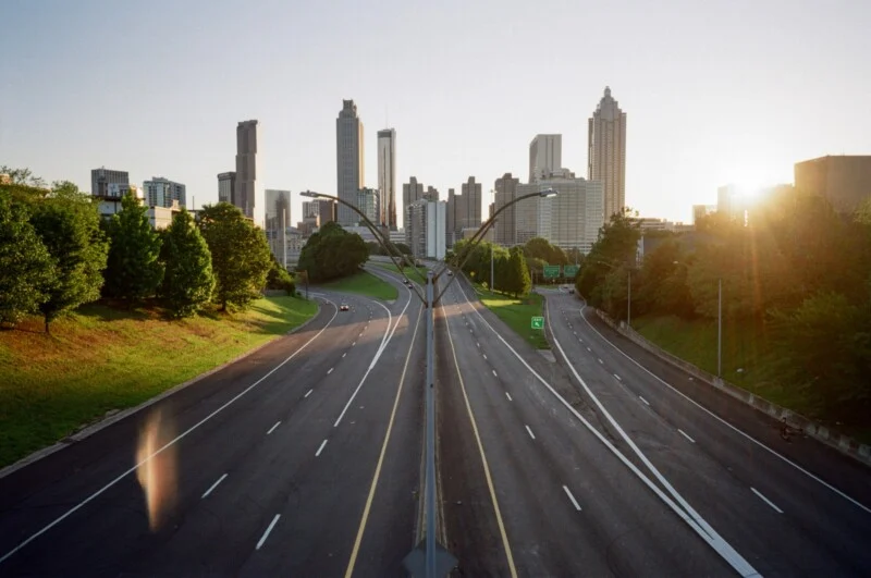 Empty highway with multiple lanes, divided by a median, leading toward a city skyline with tall buildings at sunrise or sunset. Trees line both sides of the road, and sunlight streams in from the right.