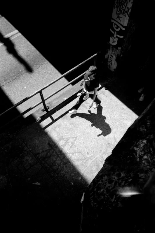 A person wearing a hat walks along a sunlit sidewalk, casting a prominent shadow. The scene is framed by strong contrasts of light and shadow, with graffiti visible on a nearby wall.