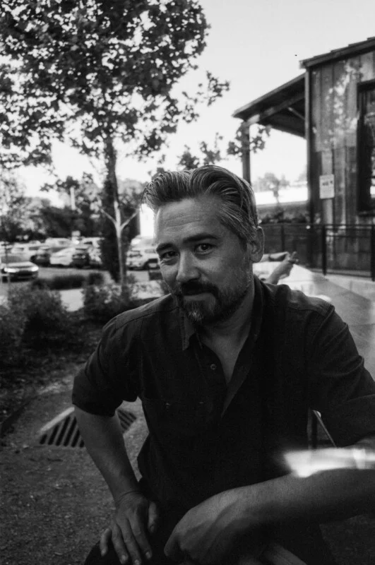 A man with short hair and a beard sits outdoors near a building, looking at the camera. Parked cars and trees are visible in the background. The image is in black and white.