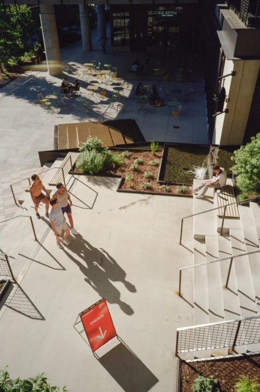 A sunny outdoor patio with people relaxing on chairs and walking. Three people walk past a red sign that says "CHECK-IN" with an arrow. Another person sits on steps near a small fountain and plants.