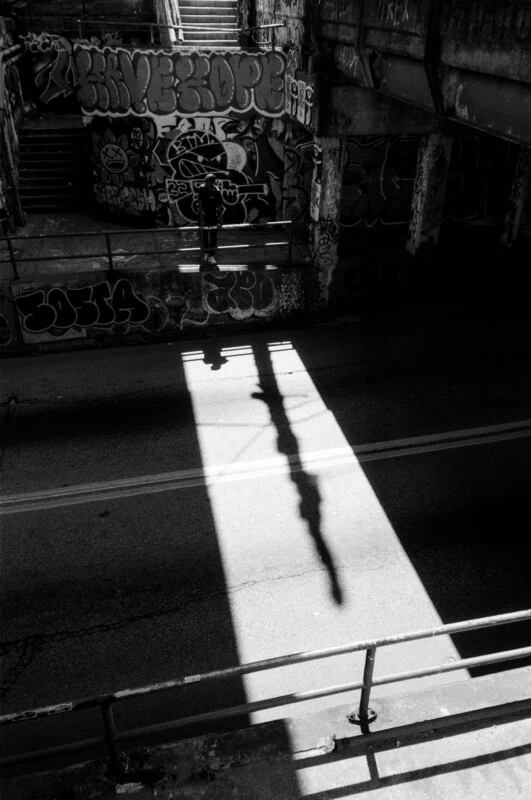 A person stands in a shadowy underpass covered in graffiti, as strong sunlight casts their silhouette onto the street below through a rectangular patch of light.