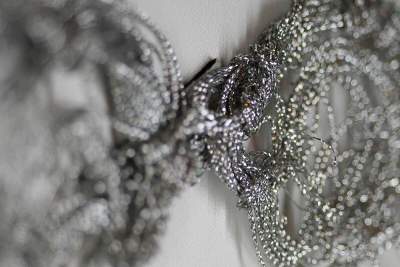Close-up of tangled silver chains hanging on a white surface, with the foreground in sharp focus and the background blurred. The intricate links create a textured, shiny pattern.