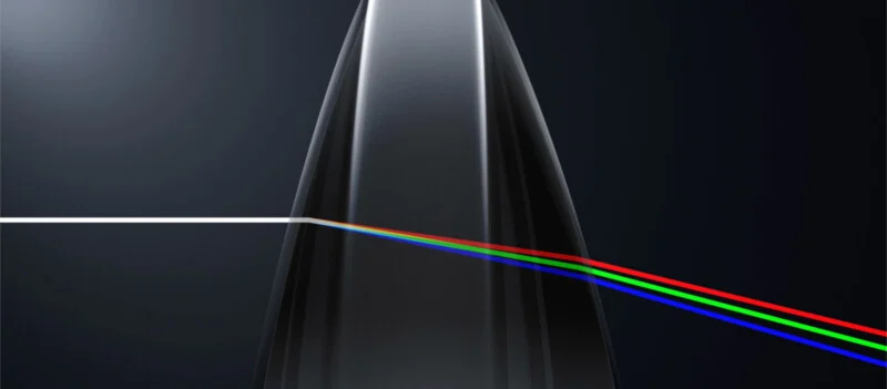 A narrow white light beam enters a transparent prism, splitting into three colored beams—red, green, and blue—on a dark background, illustrating light dispersion.