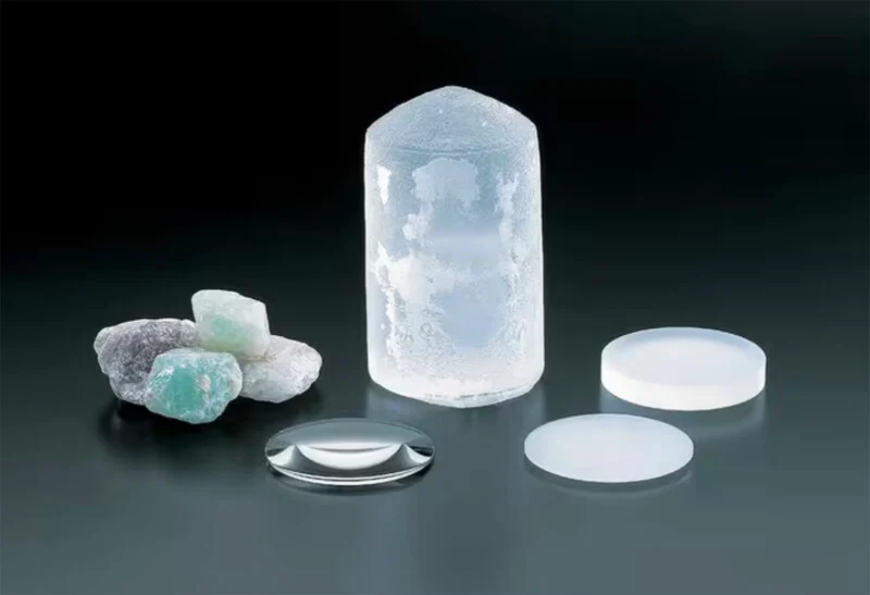 Several rough green and white crystals, a large clear synthetic crystal, and three polished optical lenses are displayed on a dark surface, illustrating the process of transforming raw minerals into finished lenses.