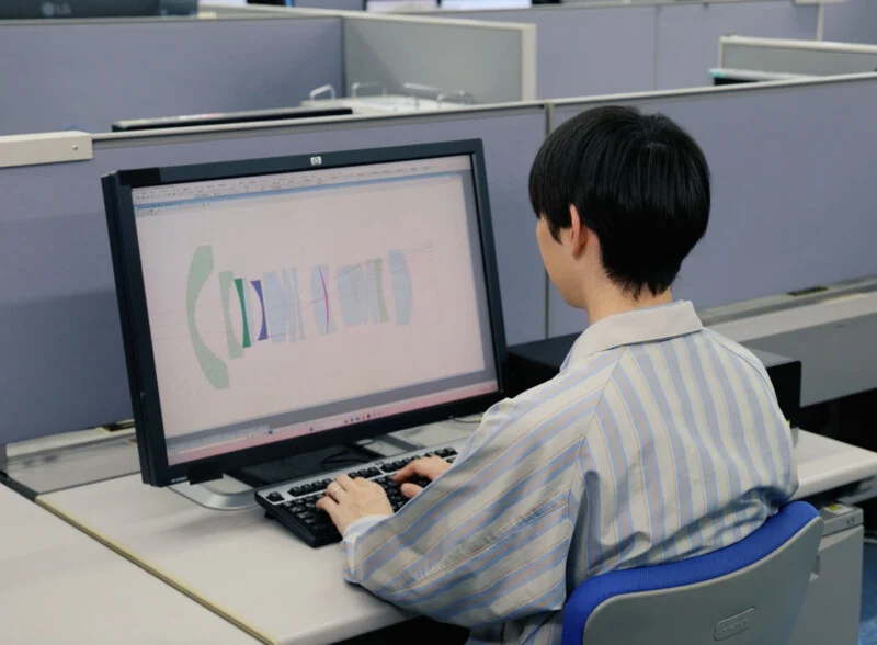 A person in a striped shirt sits at a desk in an office, working on a computer displaying colorful technical diagrams or schematics.