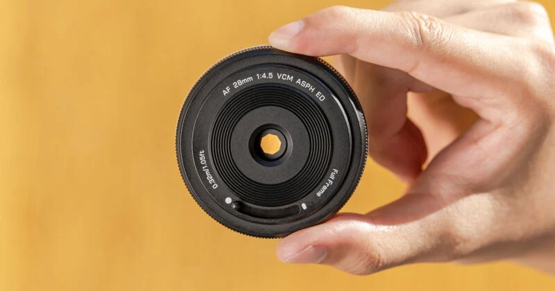 A hand holding a camera lens with text around the front rim reading "AF 28mm 1:4.5 VCM ASPH ED" against a blurred yellow background.