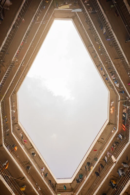Looking up from the ground floor of a multi-story building with a polygon-shaped opening, balconies lined with railings, and colorful clothes hanging from the upper floors against a bright sky.