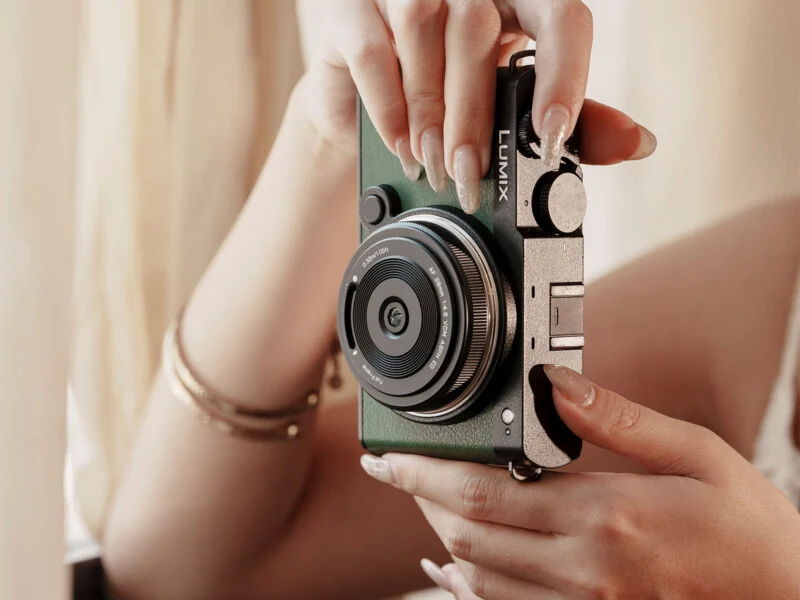 A person with manicured nails and bracelets holds a green Lumix camera with both hands, preparing to take a photo. Soft natural light enters from the side.