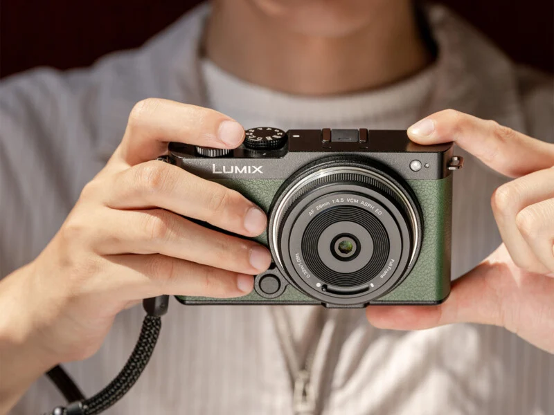 A person holds a green Lumix digital camera with both hands, preparing to take a photo. The camera is centered in the image, and the person wears a light-colored, striped shirt.