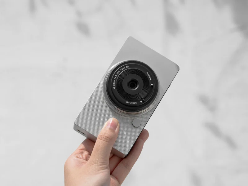 A hand holds a modern, slim digital camera with a large lens and textured grip against a soft, blurred background.