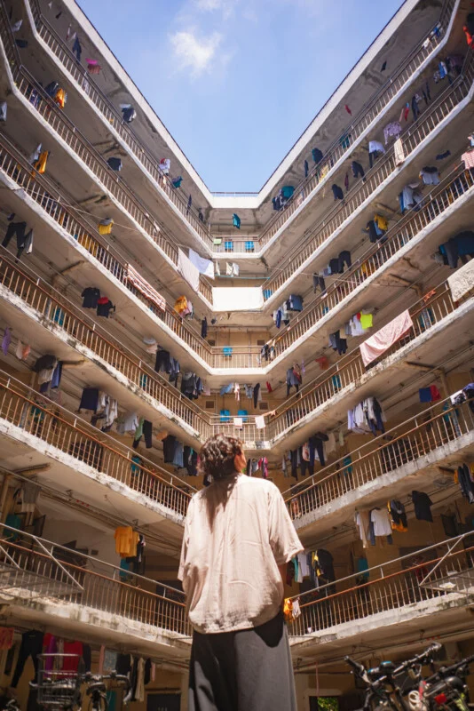 A person stands in the courtyard of a multi-story apartment building, looking up at balconies lined with laundry hanging to dry under a bright blue sky.