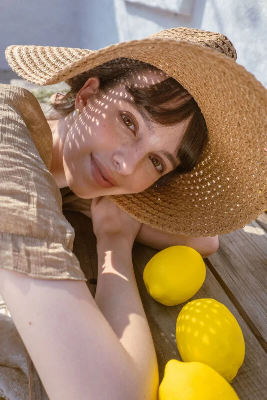 A woman wearing a large straw hat smiles while resting her arms on a wooden table with three lemons in sunlight, creating dappled light patterns on her face.
