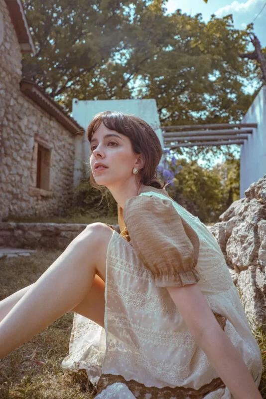 A young woman with brown hair sits outdoors on grass, wearing a light-colored dress with puff sleeves. Stone buildings and green trees surround her, and sunlight filters through the scene. She looks thoughtfully into the distance.