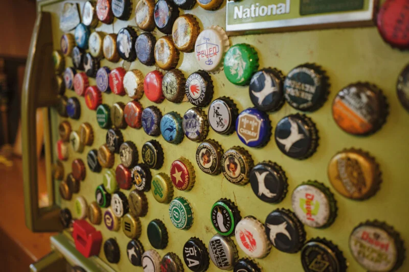 A collection of colorful bottle caps arranged in rows on a pale green, vintage metal surface, some with logos and text, creating a vibrant and eclectic display.