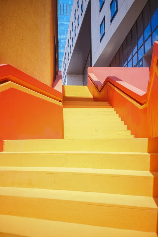 Bright yellow and orange outdoor stairs with geometric railings leading up between modern buildings, with blue sky and windows visible above.