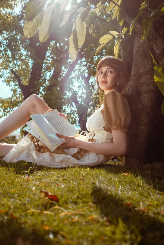 A young woman in a light dress sits on grass, leaning against a tree in sunlight, holding an open book and looking upward thoughtfully. Lush green leaves and sun rays fill the background.