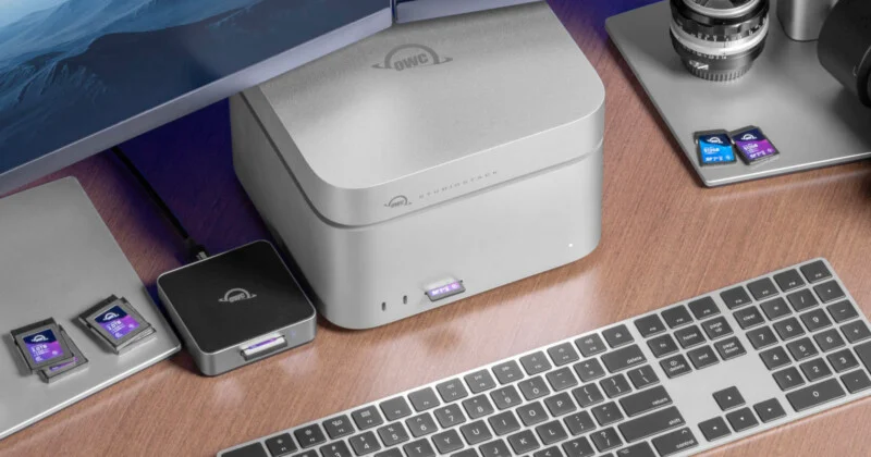 A desktop setup with a wireless keyboard, external hard drives, a camera lens, several SD cards, and a computer monitor on a wooden desk. The devices are neatly arranged beside each other.