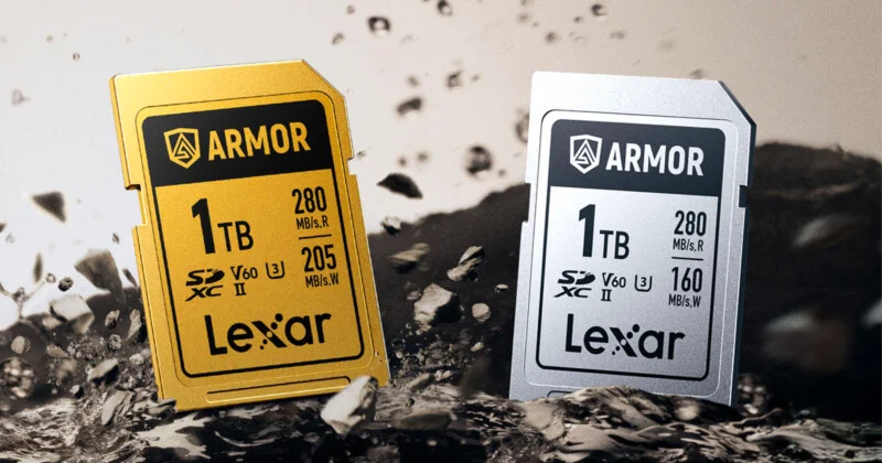 Two Lexar 1TB SD memory cards, one gold and one silver, are standing upright on a rocky surface with debris flying around them. Both cards are labeled "ARMOR" and display speed and capacity details on the front.