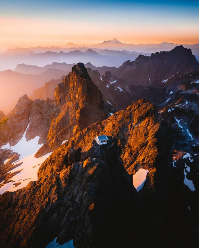 A small mountain cabin sits atop a rugged, rocky peak with patches of snow, surrounded by dramatic mountain ranges under a colorful sunrise or sunset sky.