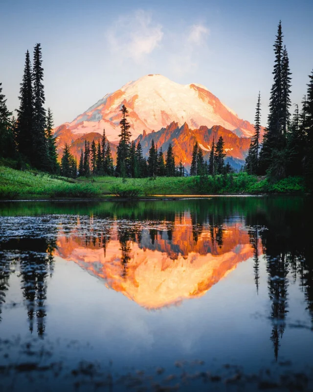 Snow-capped mountain at sunrise or sunset, glowing with orange light, reflected in a calm lake surrounded by evergreen trees and green grass under a clear sky.