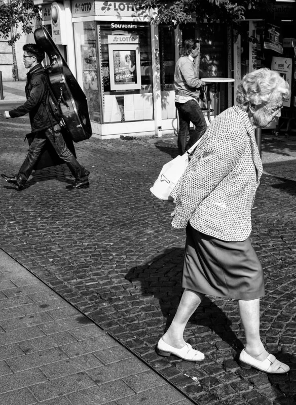 An elderly woman walks on a cobblestone street, while a man carrying a large instrument case walks behind her. Another man stands by a shop with "Souvenirs" and "Lotto" signs. The scene is in black and white.