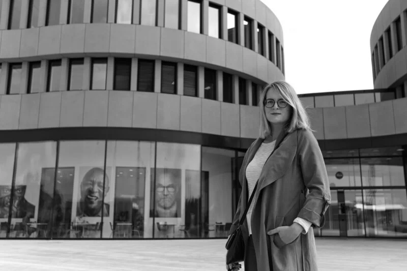 A woman wearing glasses and a long coat stands outside a modern building with large windows and round architectural features. Portraits are visible on display inside the building. The image is in black and white.