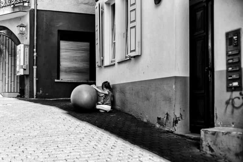 A young girl sits on a cobblestone street, leaning against a building with closed shutters, and touches a large exercise ball beside her. The scene is in black and white.