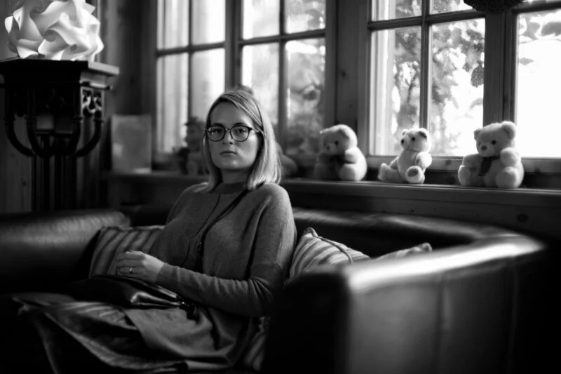 A woman with glasses sits on a couch in a cozy room, looking at the camera. Plush teddy bears are lined up on the windowsill behind her. The image is in black and white.