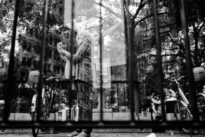 Black and white photo of a person reading a newspaper outdoors, seen through vertical metal bars. Two bicycles are parked on either side, with trees and buildings in the background.