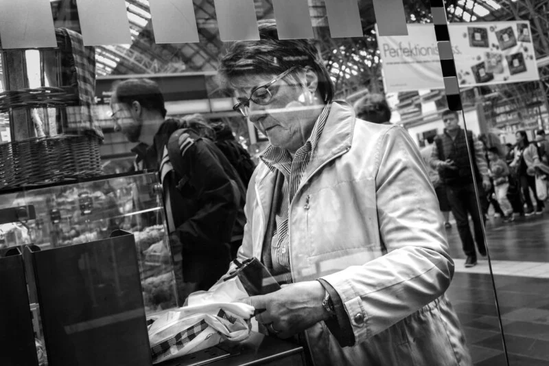A woman wearing glasses and a light coat handles a container at a market or store counter, with people walking and shopping in the busy background. The image is in black and white.
