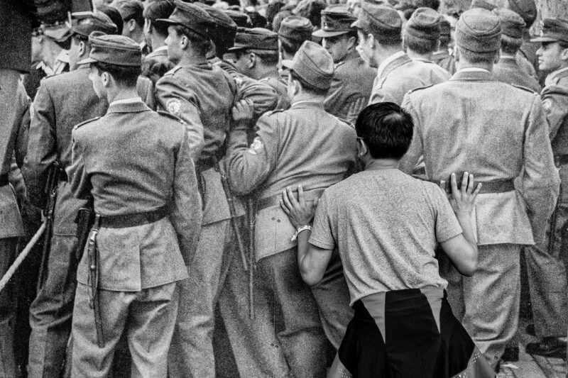 A person in a t-shirt and jacket leans against a large black-and-white photo of uniformed soldiers, pressing their hands to the image as if trying to interact with the scene.