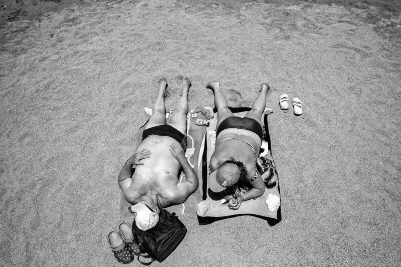 Two people lie side by side on towels at the beach, wearing swimsuits and caps. Sandals and personal items are placed near their heads on the sand. The photo is in black and white, viewed from above.