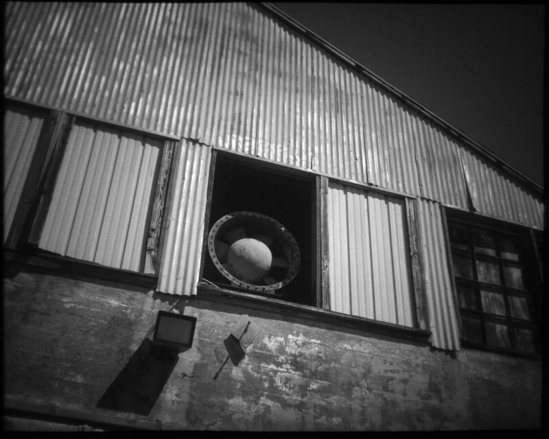 Black and white photo of an industrial building with corrugated metal siding, a large circular fan in an open window, and a security light mounted on the weathered exterior wall.