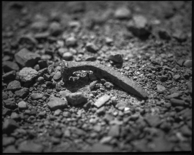 A weathered, rusty railroad spike lies among small rocks and gravel on the ground, captured in black and white with strong shadows and a rough texture.