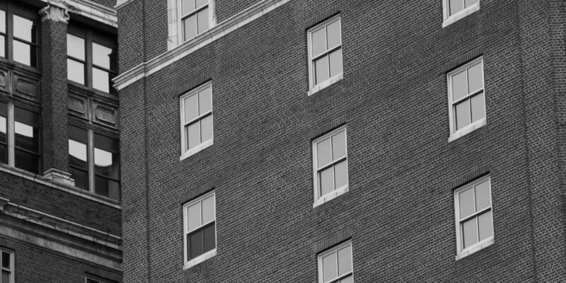 A black and white photo shows the exterior of a brick building with several windows, some open and some closed, alongside the corner of another older building with decorative trim and large, divided windows.
