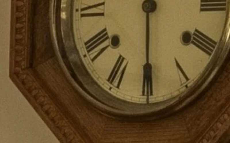 A close-up of a wooden wall clock showing Roman numerals, with the time reading approximately 6:00. The clock face is partially visible, and the wood frame has a carved pattern along the edge.