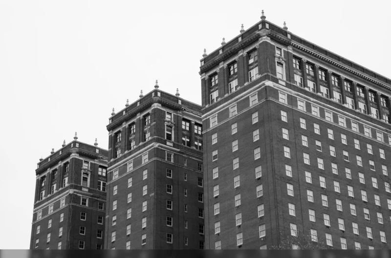 Black and white photo of three adjoining multi-story brick buildings with decorative rooflines and many windows, viewed from a low angle against a plain sky.