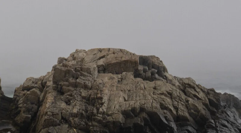 A large, rugged rock formation sits by the ocean under a gray, foggy sky, with faint waves visible in the background.