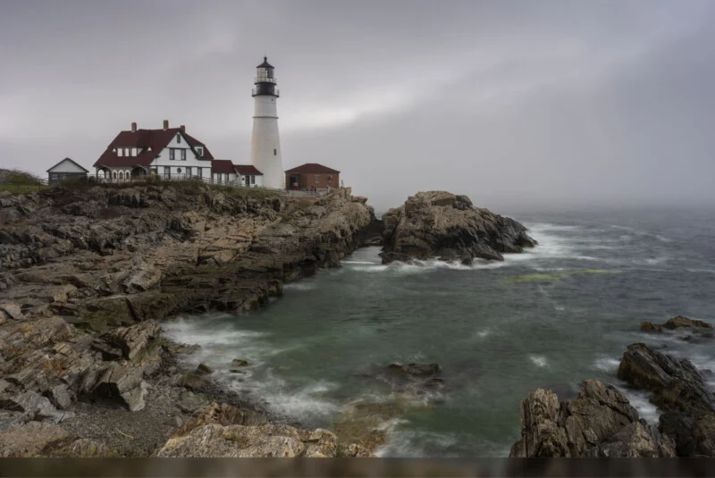 A lighthouse and adjacent buildings sit atop rocky cliffs overlooking a turbulent, misty sea under a cloudy sky. Waves crash against the rocks, and the scene appears calm yet dramatic.