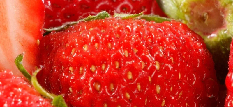 Close-up of a ripe, red strawberry showing its textured surface, small yellow seeds, and green leafy top, with sections of other strawberries visible around the edges.