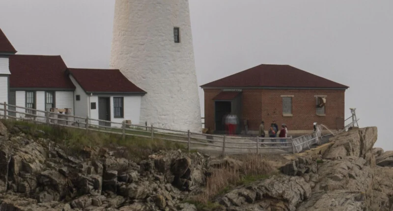 A lighthouse with a white tower and nearby brick and white buildings sits on rocky terrain, with several people standing near the entrance on a cloudy day.