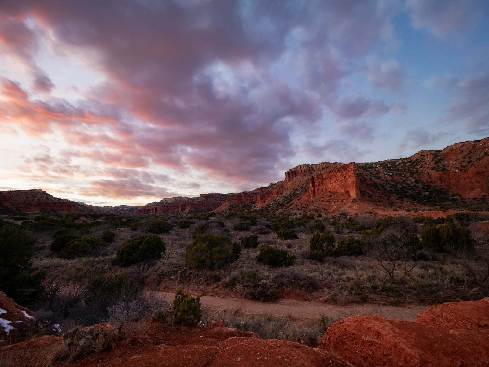 Red rock formations and green shrubs under a dramatic, colorful sunset sky with clouds, in a rugged, semi-arid landscape. A dirt path winds through the foreground.