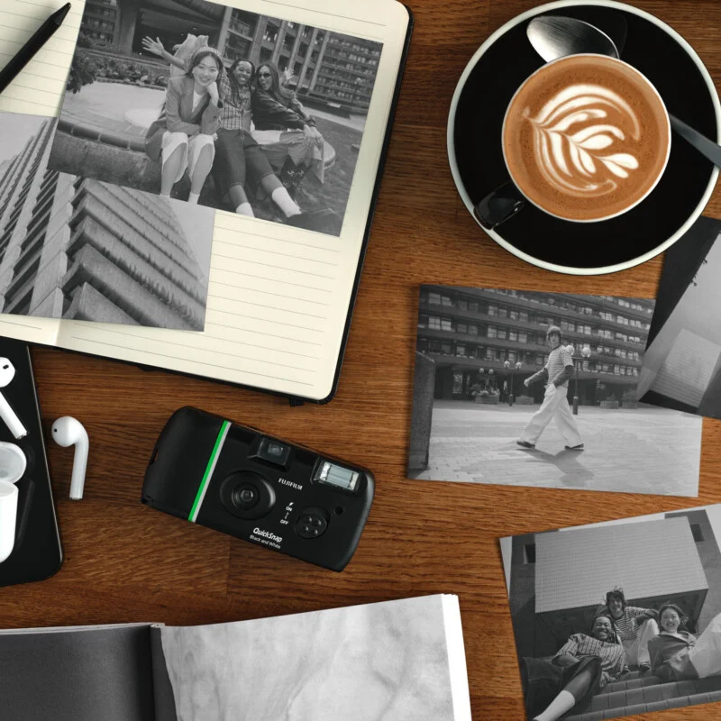 A wooden table with black-and-white photos, an instant camera, a notebook, earbuds, and a cup of latte art coffee. Some photos show people posing together and walking in an urban setting.