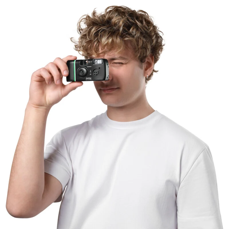 A person with curly hair wearing a plain white t-shirt is holding a disposable camera up to their eye, as if taking a photo, against a white background.