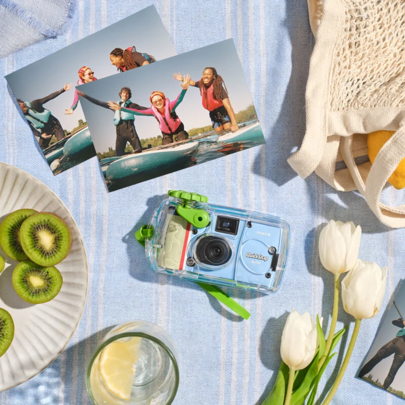 A blue waterproof camera, two surf photos, a plate of sliced kiwi, a glass of lemon water, white tulips, and a beige bag are arranged on a striped light blue cloth.