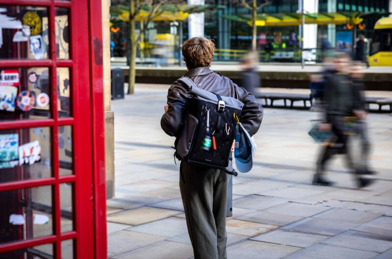 A person with short hair and a leather jacket walks on a city sidewalk, carrying a backpack with a water bottle. A red phone booth with stickers is on the left, and blurred pedestrians and a yellow tram are in the background.