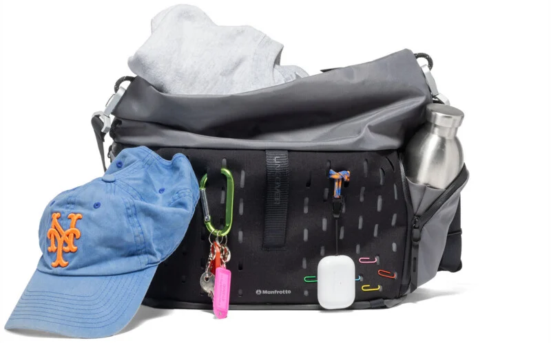 A black and gray shoulder bag with a water bottle in a side pocket, a sweatshirt inside, a blue New York Mets cap, colorful keychains, carabiners, AirPods case, and paperclips attached to the front.